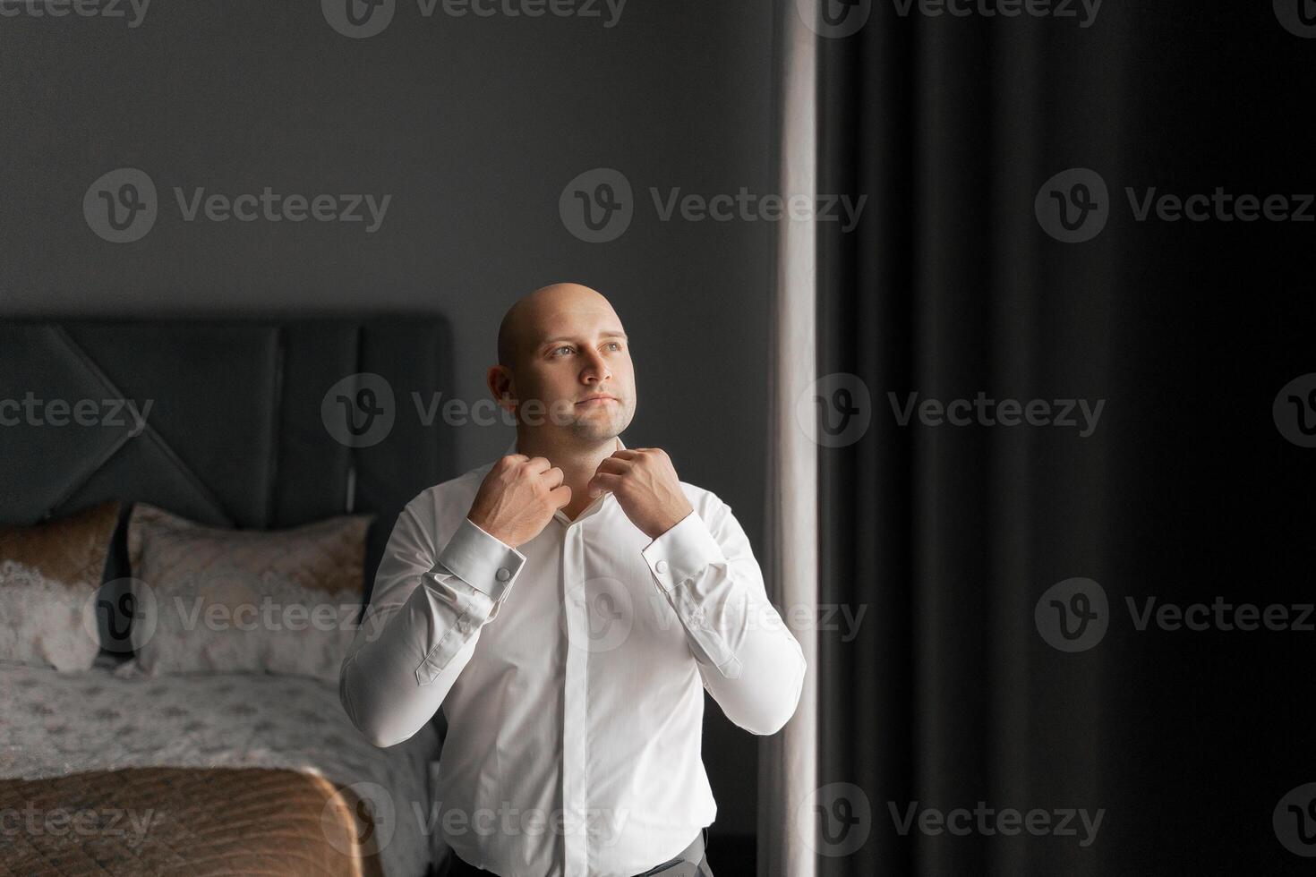 A man in a white shirt is getting ready to tie his tie. He is standing in front of a window, and the room is dimly lit photo