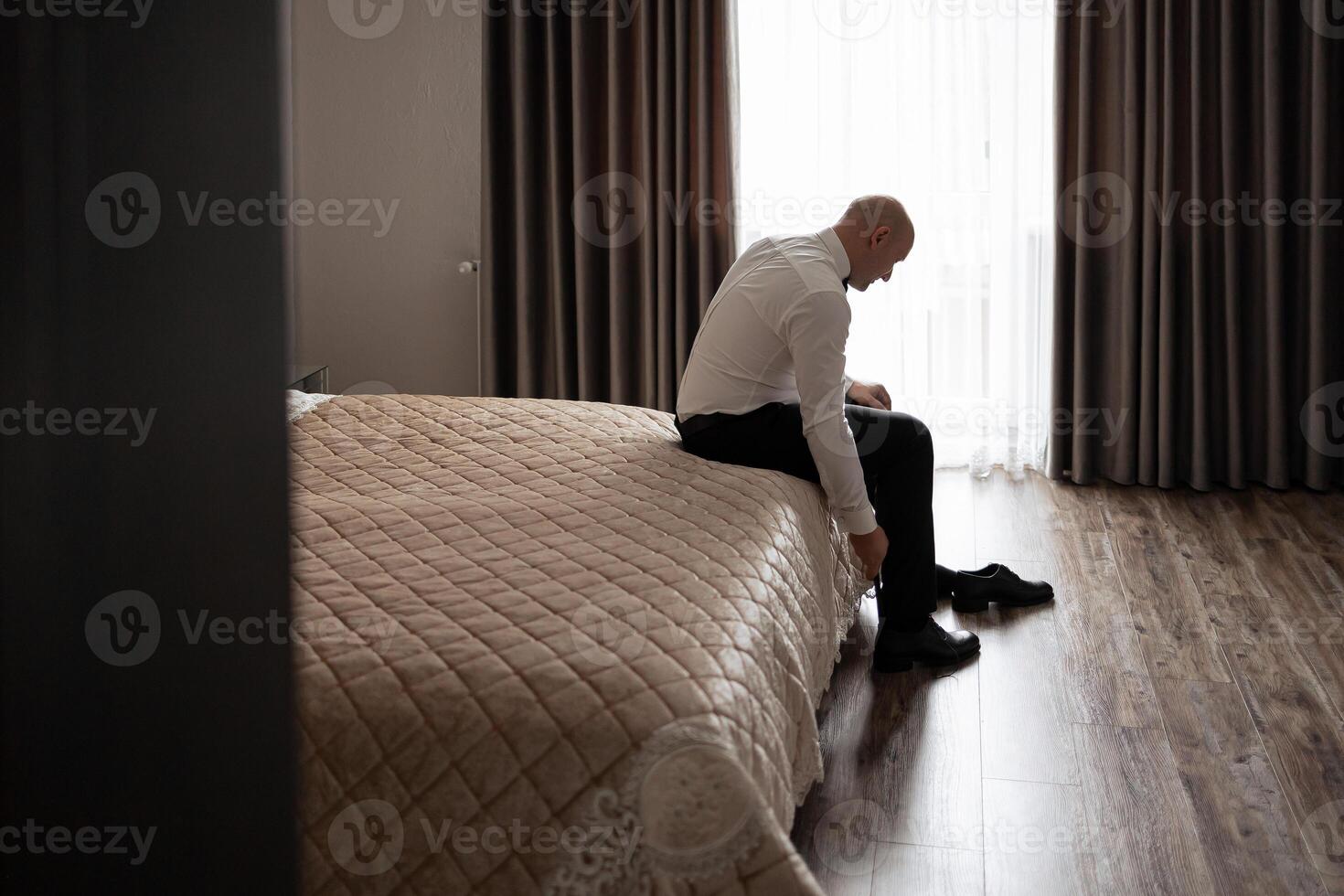 A man in a suit is sitting on a bed in a hotel room. He is adjusting his tie and shoes photo
