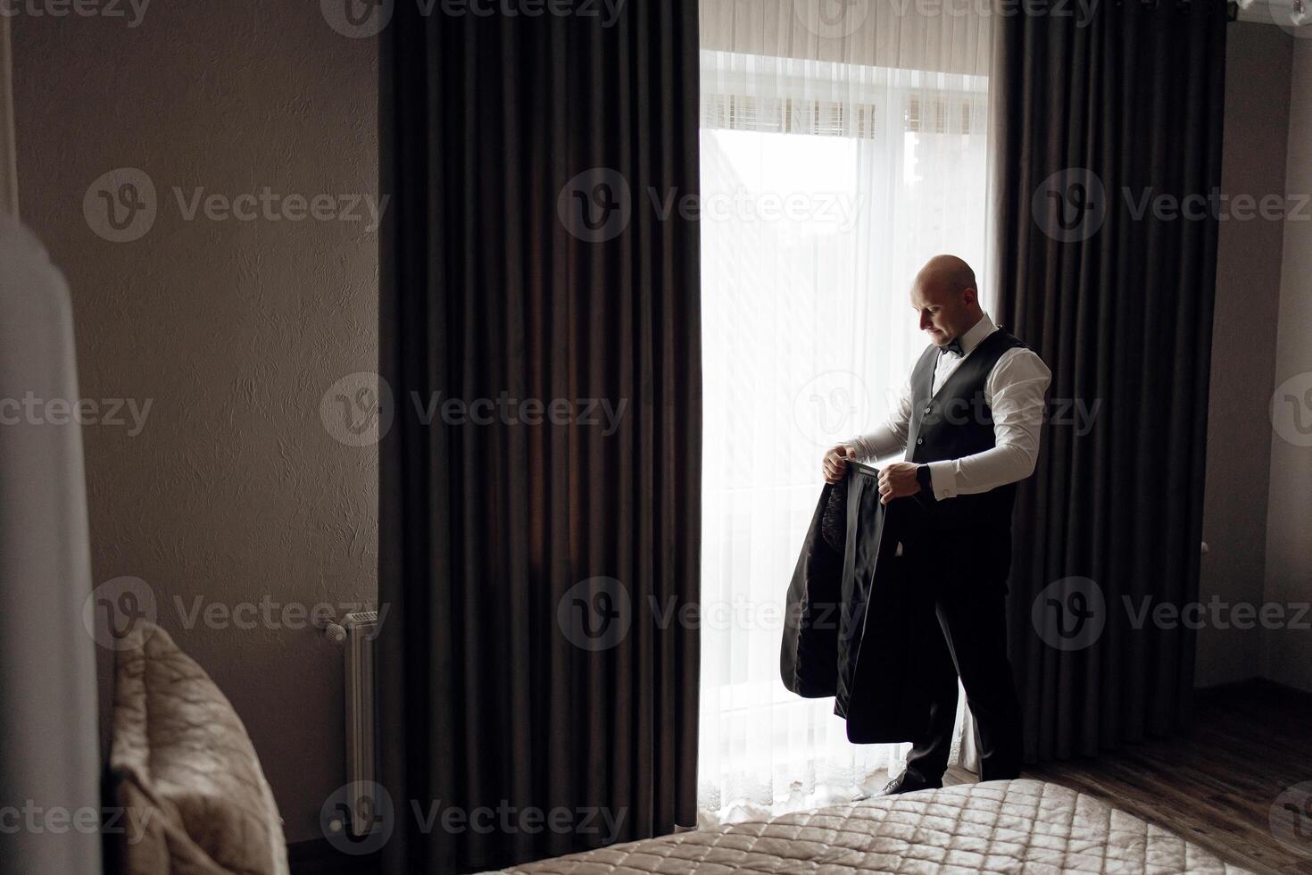 A man in a suit is standing in front of a window, getting dressed. The room is dimly lit, and the curtains are drawn, creating a sense of privacy and intimacy photo
