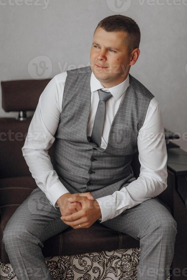 A man in a gray vest and white shirt sits on a brown chair. He is wearing a gray tie and has his hands on his knees. Concept of formality and professionalism photo