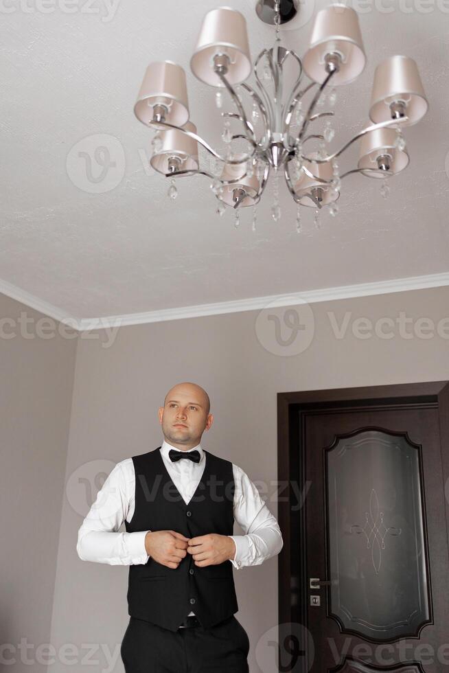 A man in a suit and vest stands in front of a chandelier. He is adjusting his tie and looking up at the ceiling. Scene is formal and elegant, as the man is dressed in a suit and vest photo