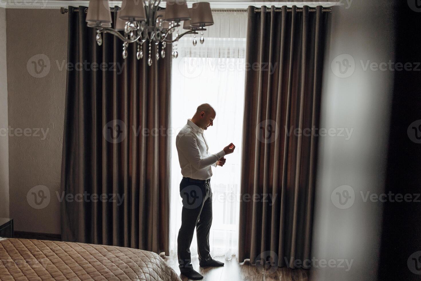 A man is standing in front of a window, adjusting his tie. The room is dimly lit, and the curtains are drawn, creating a sense of privacy and intimacy. The man is in a focused and serious mood photo