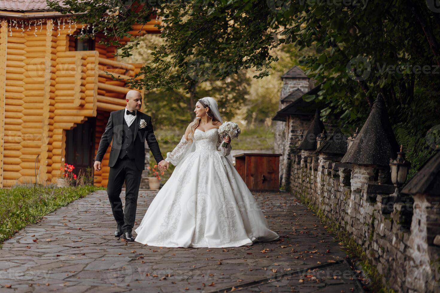 A bride and groom are walking down a path in front of a wooden building. The bride is wearing a white dress and the groom is wearing a black suit. Scene is romantic and happy photo
