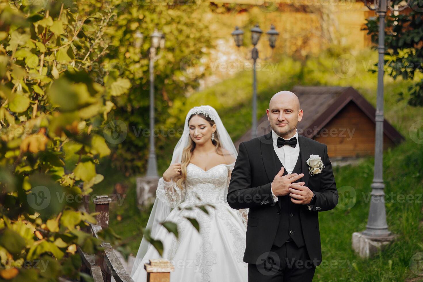 A bride and groom are walking down a path in a garden. The bride is wearing a white dress and the groom is wearing a black suit. Scene is happy and romantic, as the couple is getting married photo