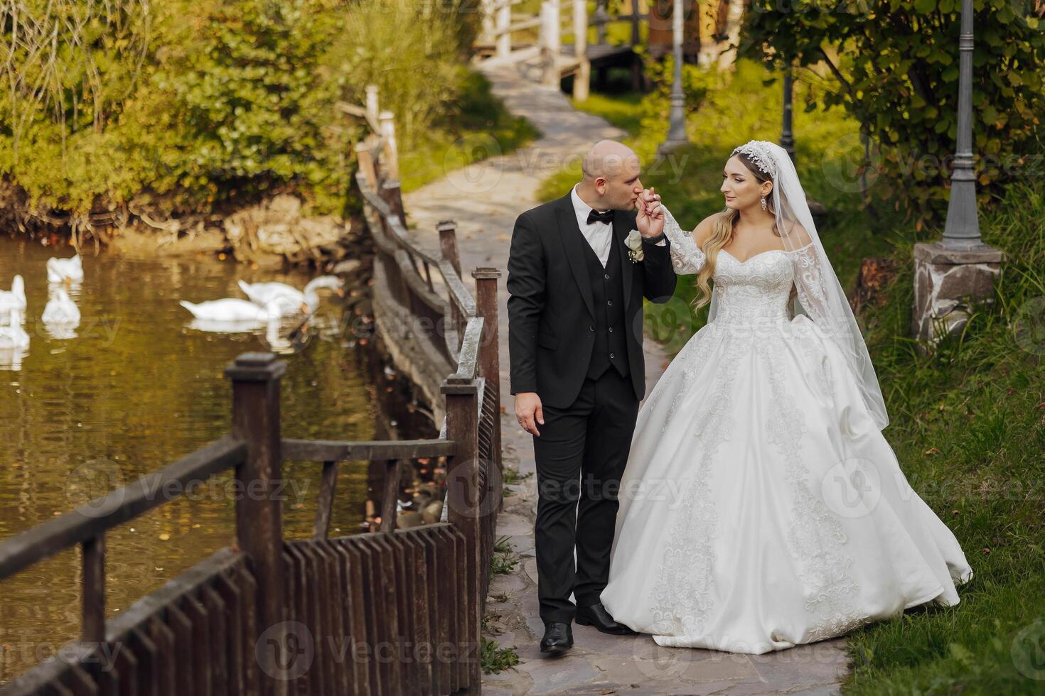A bride and groom walk along a path near a pond. The bride is wearing a white dress and the groom is wearing a black suit. There are several ducks in the pond photo