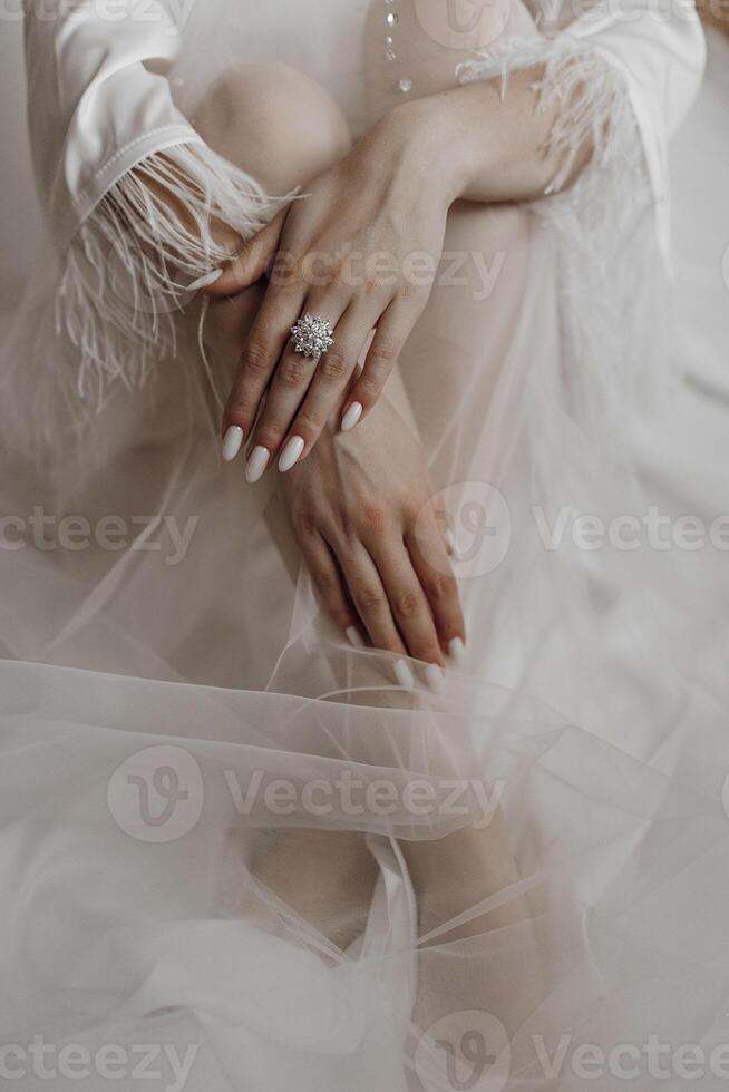A woman is sitting on a bed with a white dress and a ring on her finger. The dress is made of a sheer material, and the woman's hands are placed on her lap. Concept of elegance and sophistication photo