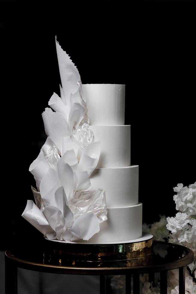 A white cake with a white leafy design on it. The cake is four layers and is placed on a table photo