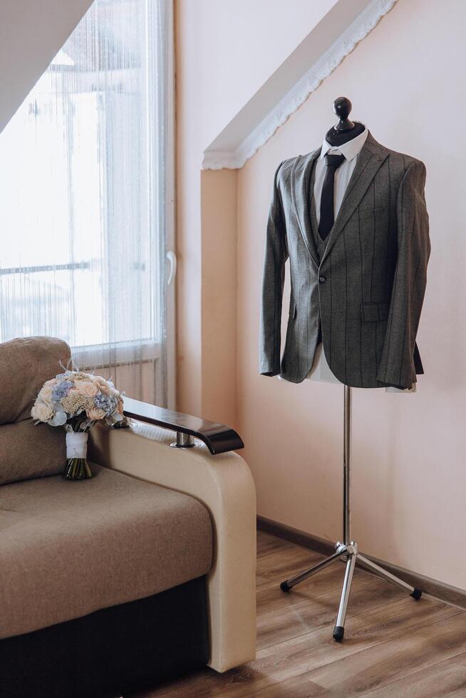 A mannequin wearing a gray suit and a black tie stands in front of a couch. A bouquet of flowers is placed on the couch photo