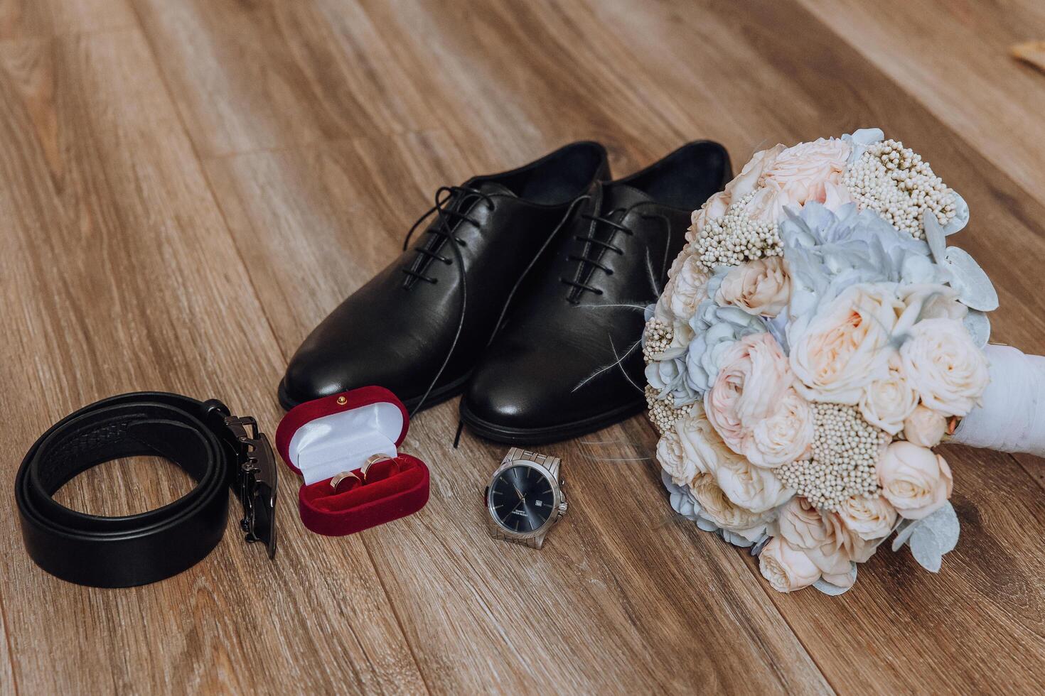 A man's shoes and a watch are displayed on a wooden floor. A bouquet of flowers is placed next to the shoes photo