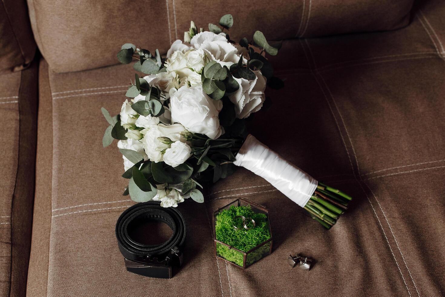 A bouquet of white flowers sits on a couch next to a belt and a small box. The flowers are arranged in a way that they look like they are about to be given as a gift photo