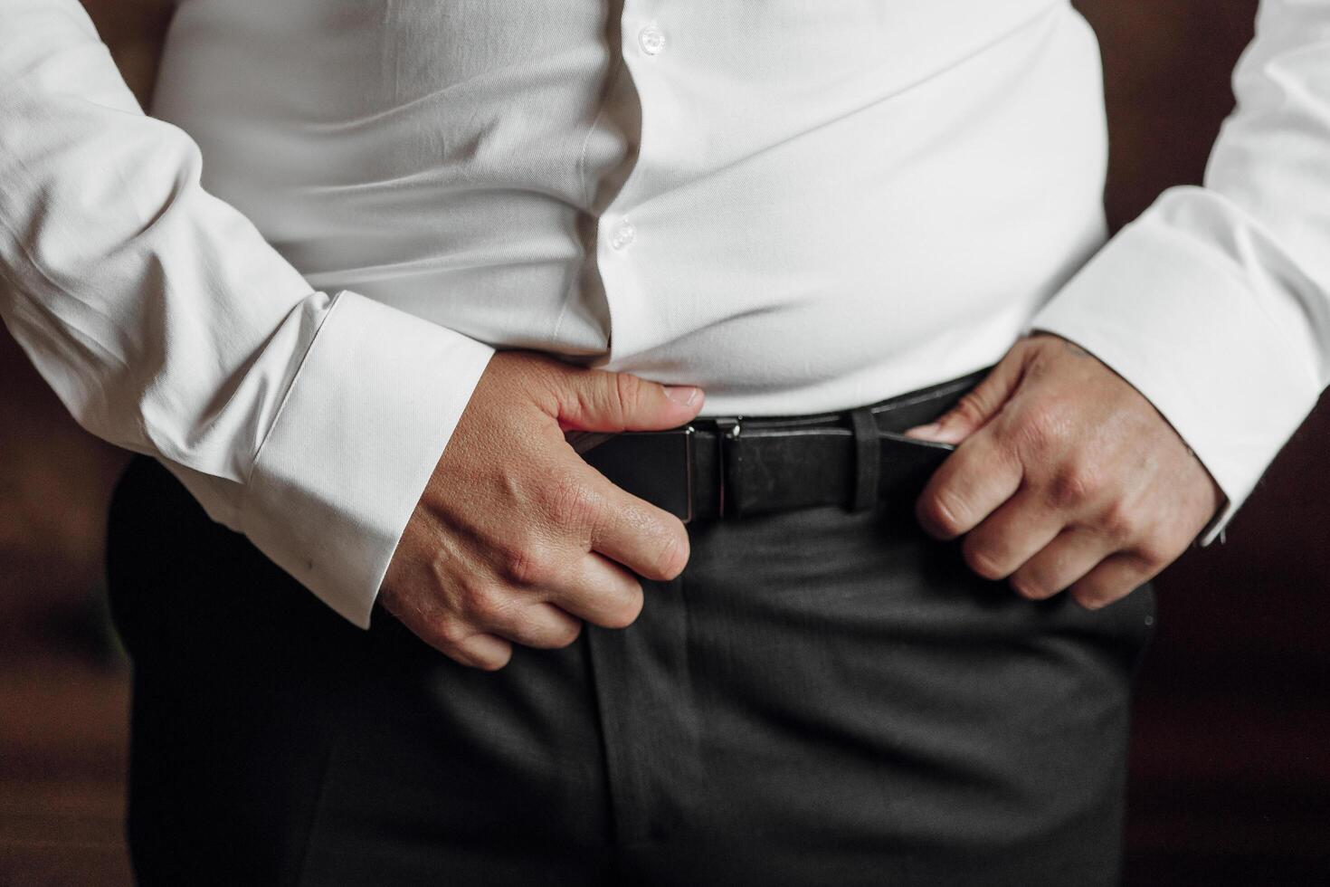A man in a white shirt and black pants is wearing a belt. He is standing with his hands in his pockets photo