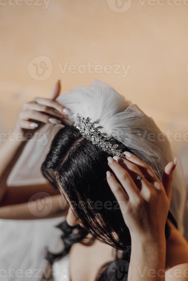 A woman is putting on a tiara with a veil. The tiara is made of gold and has a diamond in the center. The woman is wearing a white dress and has her hands on her head photo