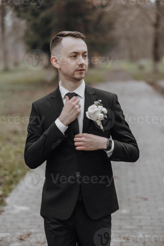 A man in a suit is wearing a white flower in his lapel. He is standing on a brick walkway photo