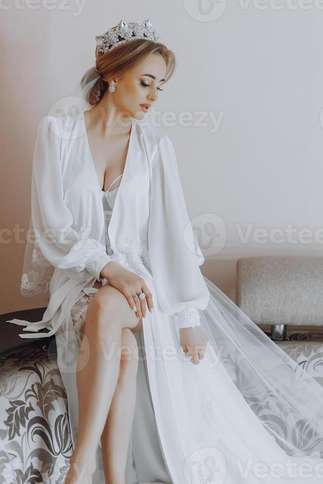 A woman in a white gown sits on a bed. She is wearing a tiara and has her legs crossed photo
