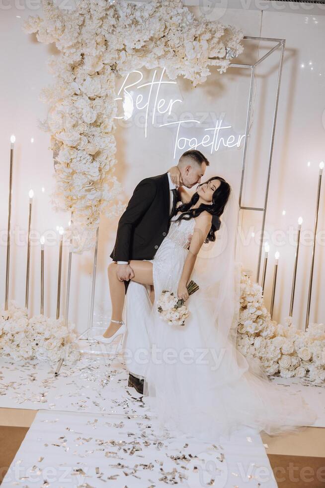A bride and groom kiss in front of a white backdrop . The bride is holding a bouquet and the groom is holding a wine glass photo
