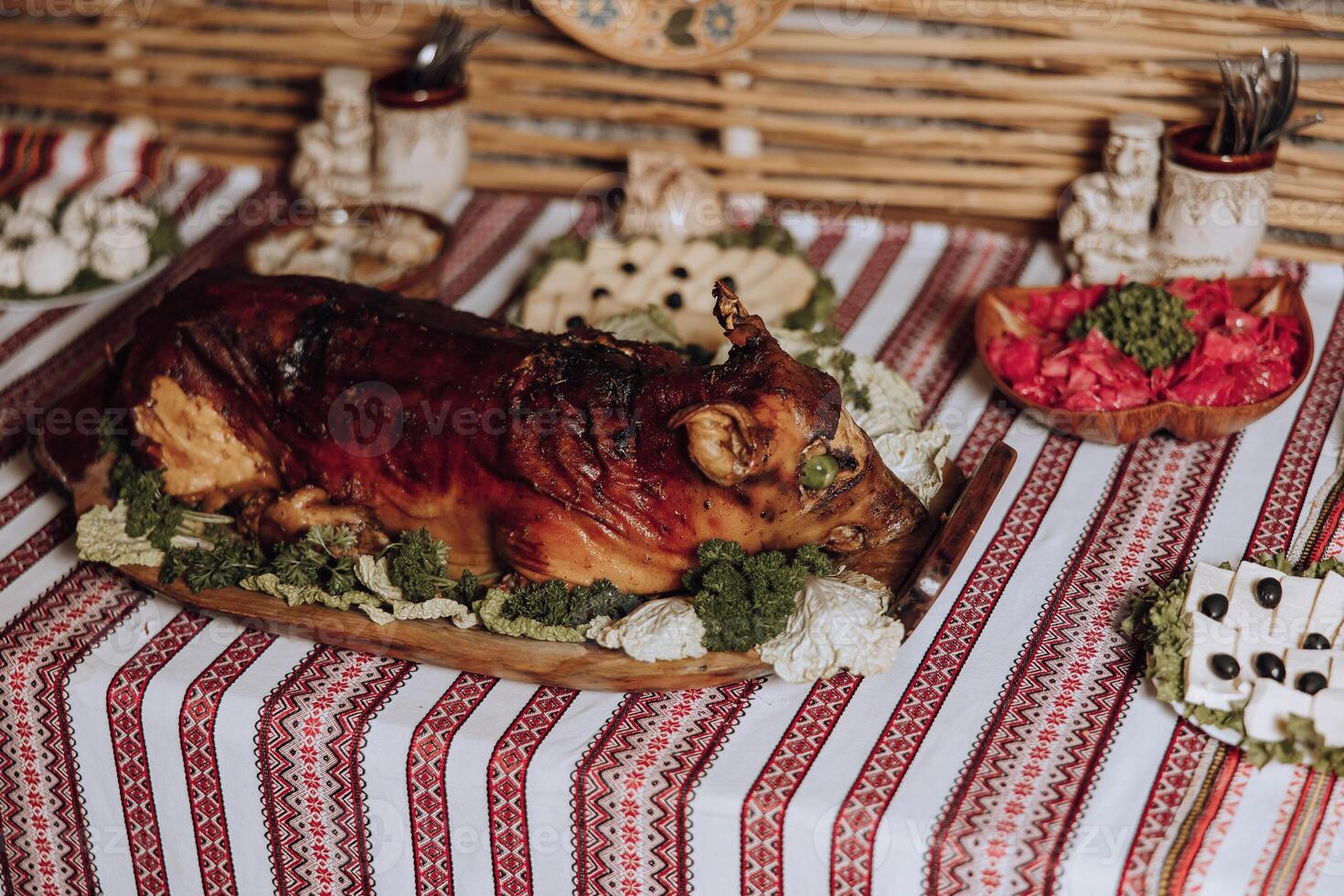 A large roast pig is on a wooden platter on a table with other food items. The tablecloth is red and white striped photo