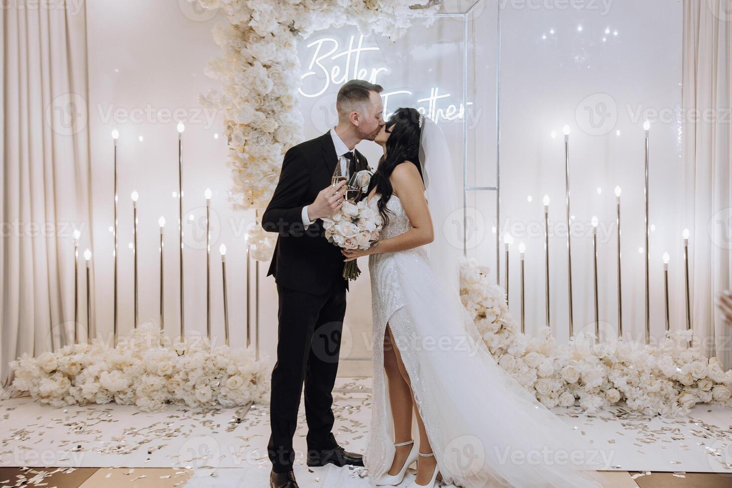 A bride and groom kiss in front of a white backdrop . The bride is holding a bouquet and the groom is holding a wine glass photo