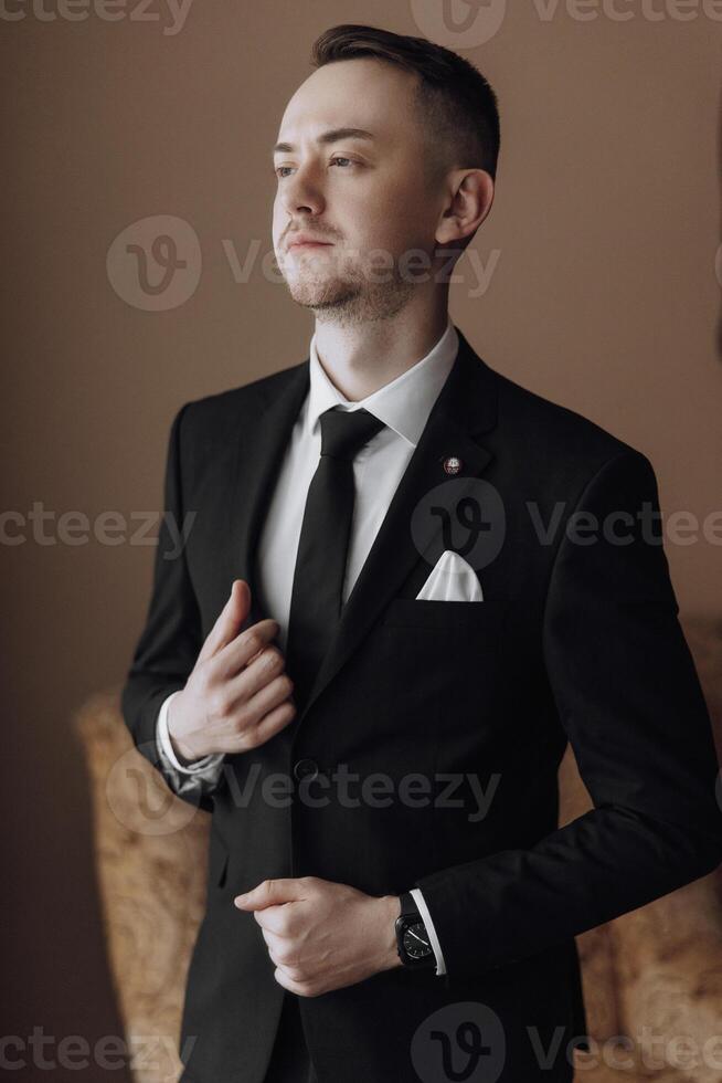 A man in a black suit and white shirt is standing in front of a chair. He is wearing a black tie and a white pocket square. The man is getting ready for a formal event or a professional occasion photo
