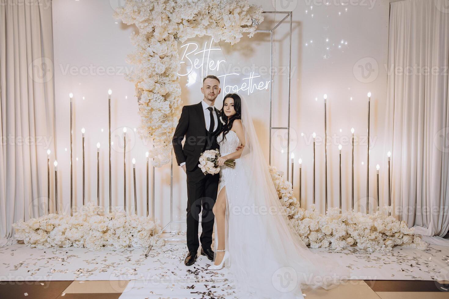 A bride and groom kiss in front of a white backdrop . The bride is holding a bouquet and the groom is holding a wine glass photo