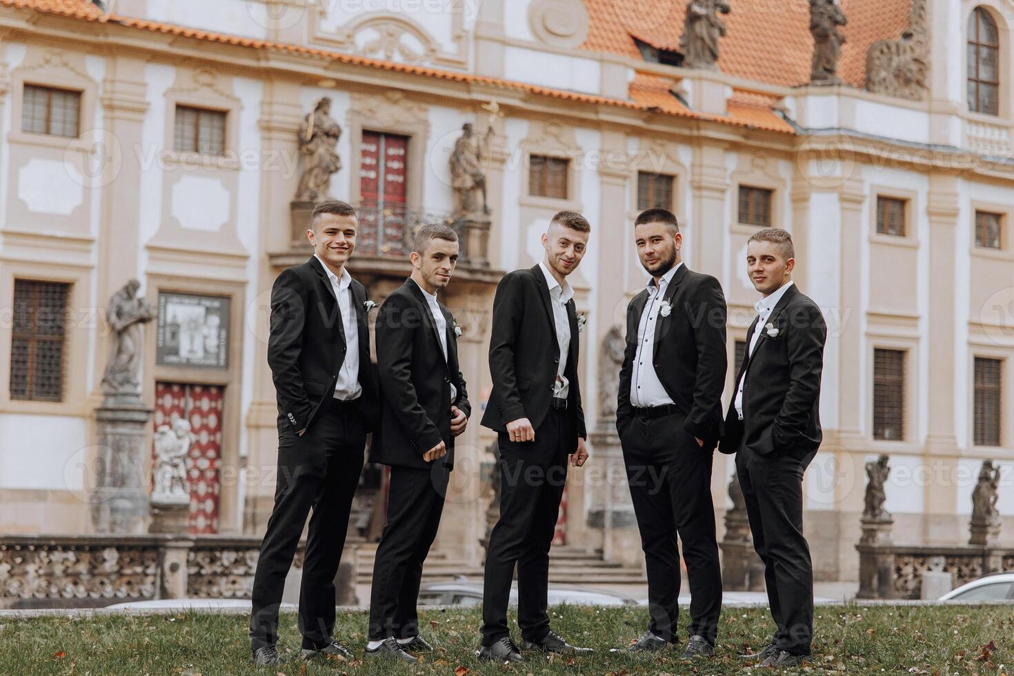A group of five men are standing in front of a large building, dressed in black suits and ties. Scene is formal and elegant, as the men are posing for a photo in their suits