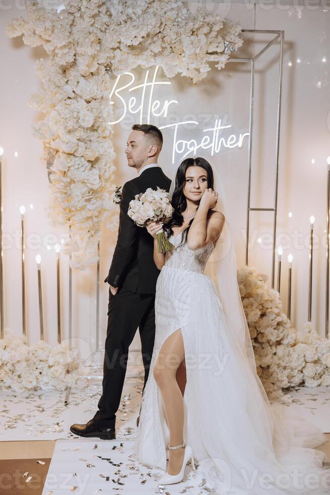 A bride and groom kiss in front of a white backdrop . The bride is holding a bouquet and the groom is holding a wine glass photo