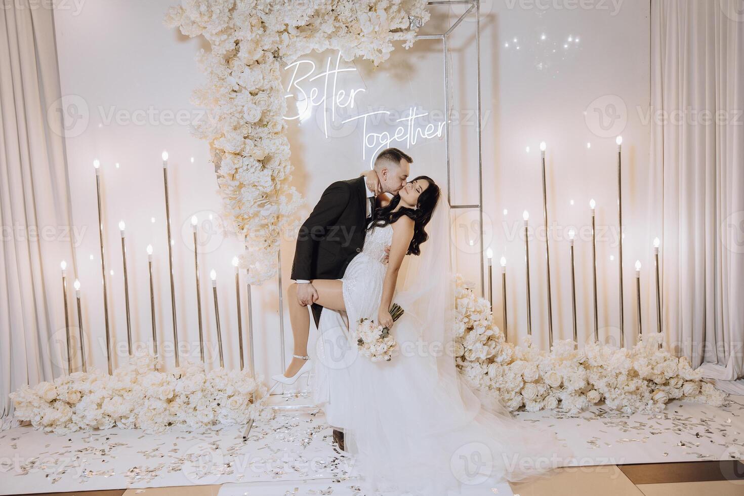 A bride and groom kiss in front of a white backdrop . The bride is holding a bouquet and the groom is holding a wine glass photo
