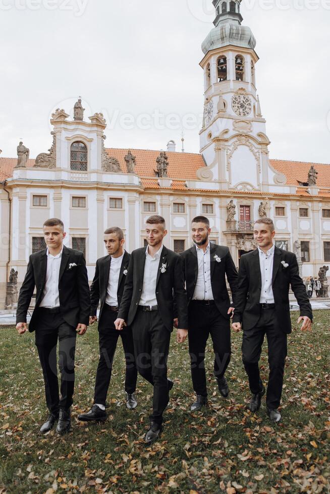 A group of five men in suits are walking together in front of a large building. The men are dressed in black and white, and they appear to be posing for a photo. Scene is formal and elegant photo