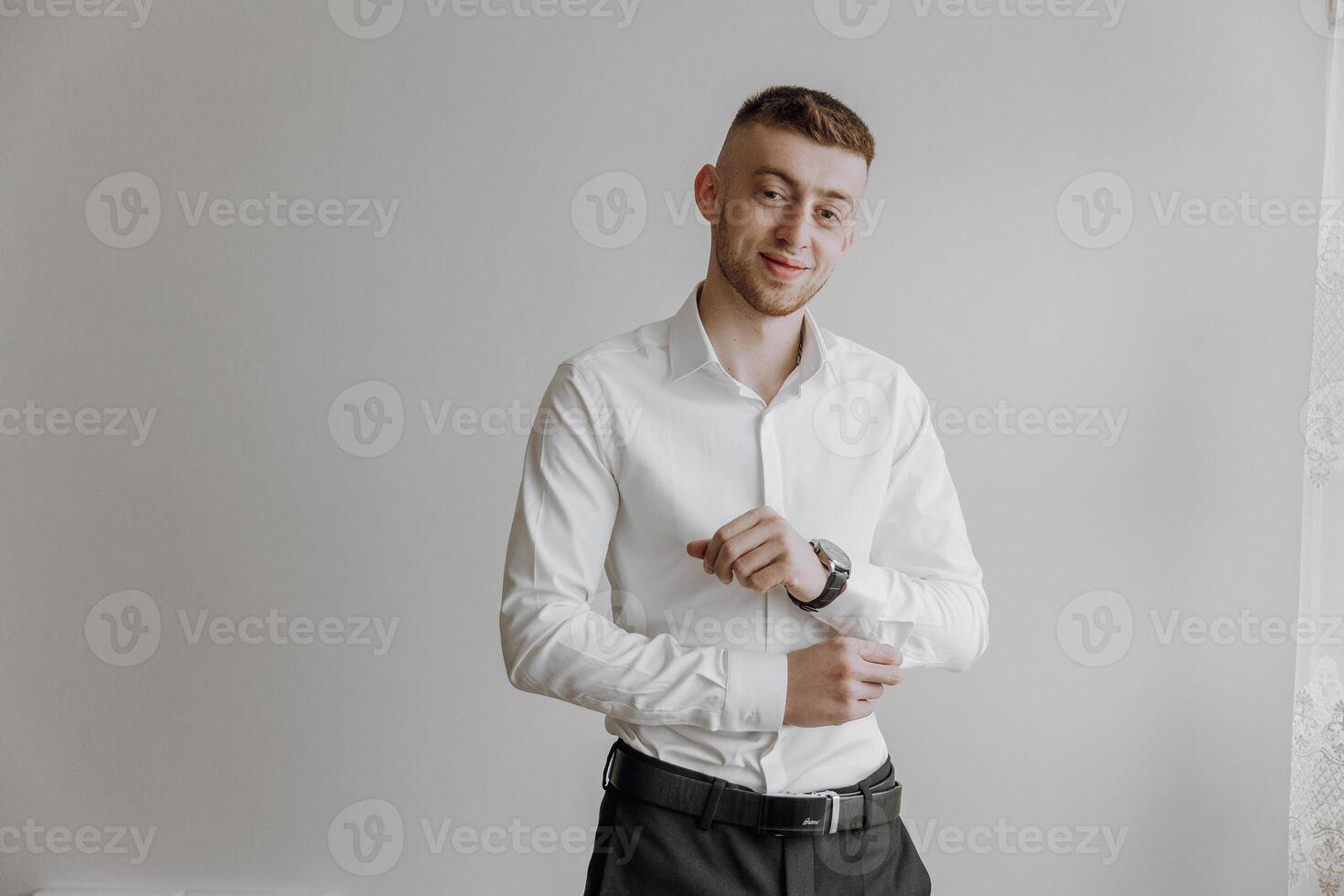 A man in a white shirt and black pants is standing in front of a white wall. He is wearing a watch and is adjusting his shirt. Concept of formality and attention to detail photo