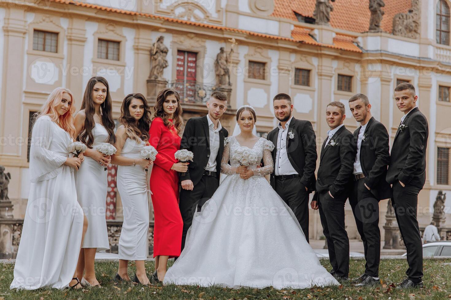 A group of people are posing for a picture in front of a building. The bride and groom are in the center of the group, with the bride wearing a white dress and the groom wearing a suit photo