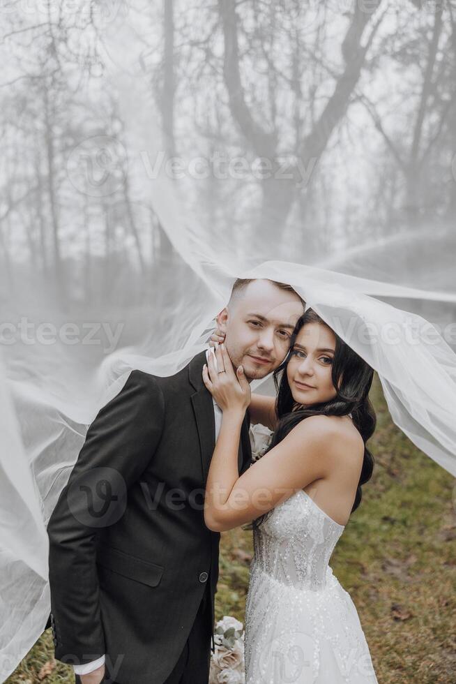 A man and woman are posing for a picture under a white veil. The man is wearing a suit and tie, while the woman is wearing a white dress. Scene is romantic and intimate photo