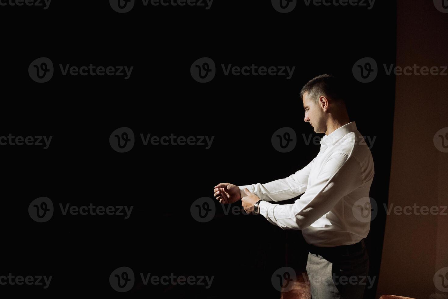 A man in a white shirt is standing in a dark room, looking at his watch. He is in a hurry and is checking the time frequently photo