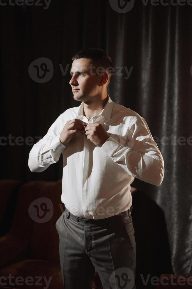 A man in a white shirt is getting dressed. He is standing in front of a curtain. The man is wearing a gray suit and gray pants photo