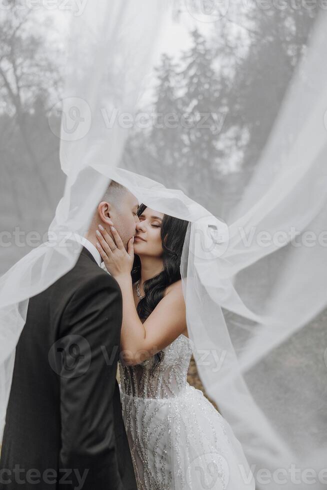 A bride and groom are kissing in front of a white veil. The bride is wearing a white dress and the groom is wearing a black suit. The scene is intimate and romantic, capturing the love photo