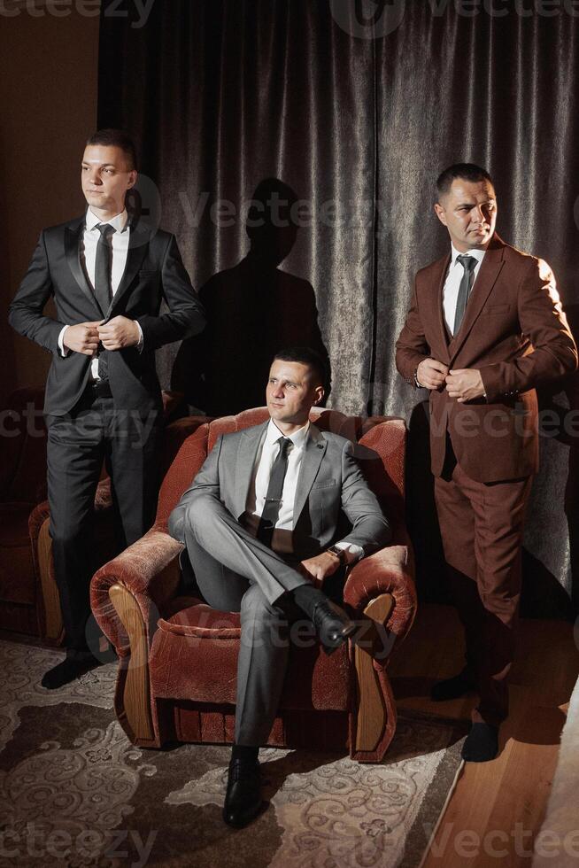 Three men in suits are posing for a photo in front of a red chair. Scene is formal and elegant, as the men are dressed in suits and ties