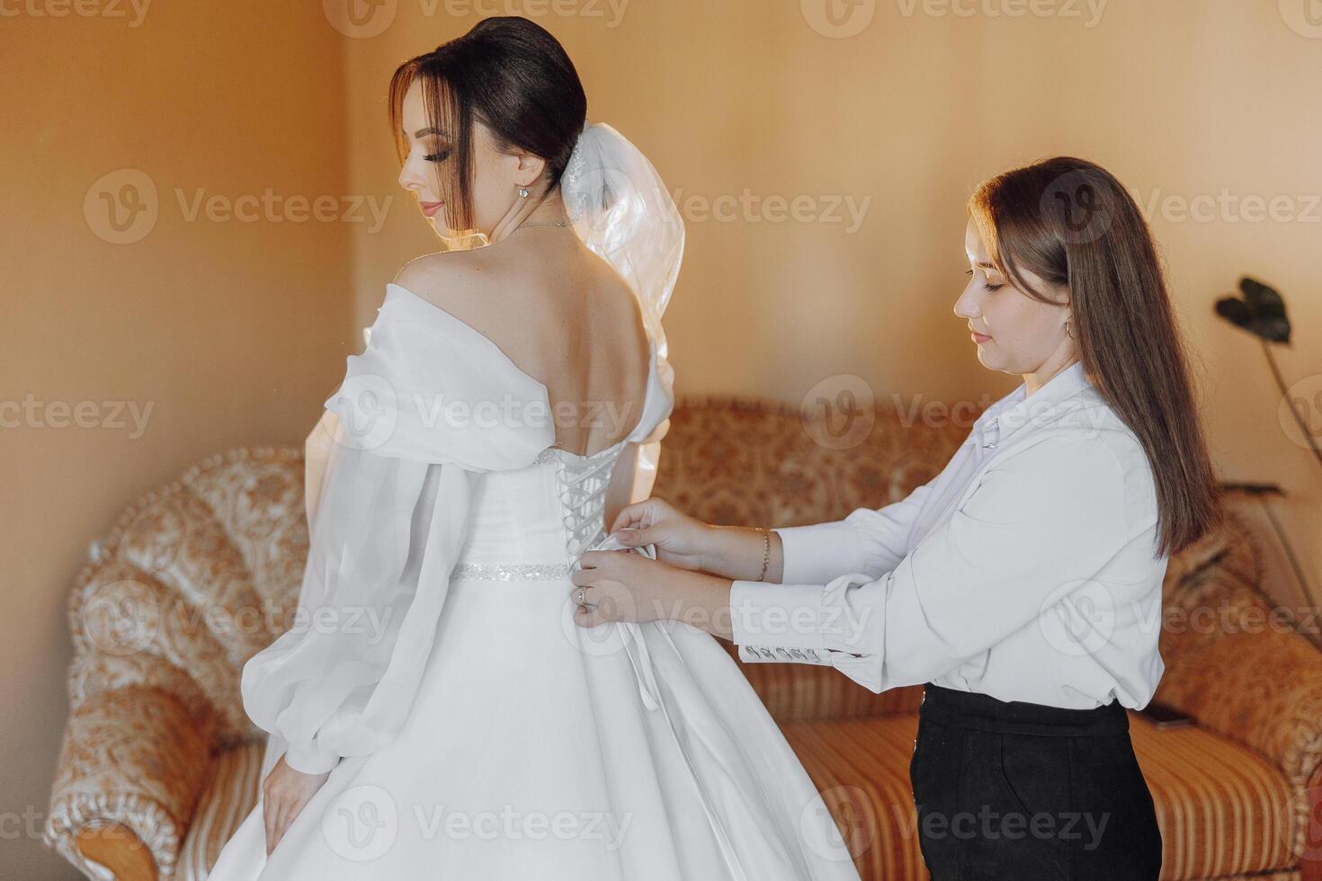 A woman is getting ready for a wedding and is being helped by another woman. The bride is wearing a white dress and the other woman is adjusting her dress. Scene is one of support photo
