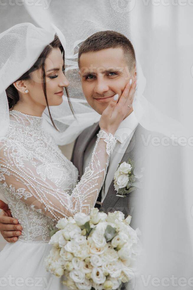 A bride and groom are embracing each other with a white veil over the bride's head. The bride is holding a bouquet of flowers and the groom is holding her hand. The scene is a wedding photo