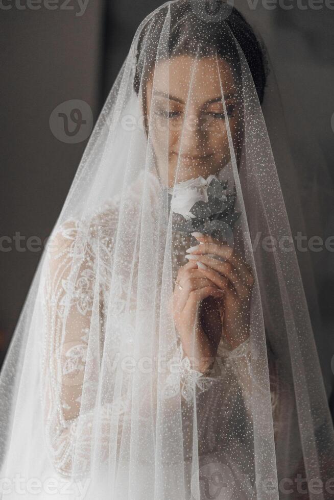 A bride is wearing a veil and holding a flower. The veil is made of a net-like material and covers her face photo