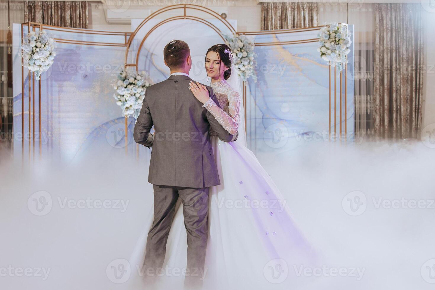 A bride and groom are dancing in front of a blue archway. The bride is wearing a white dress and the groom is wearing a gray suit. The atmosphere is romantic and joyful photo