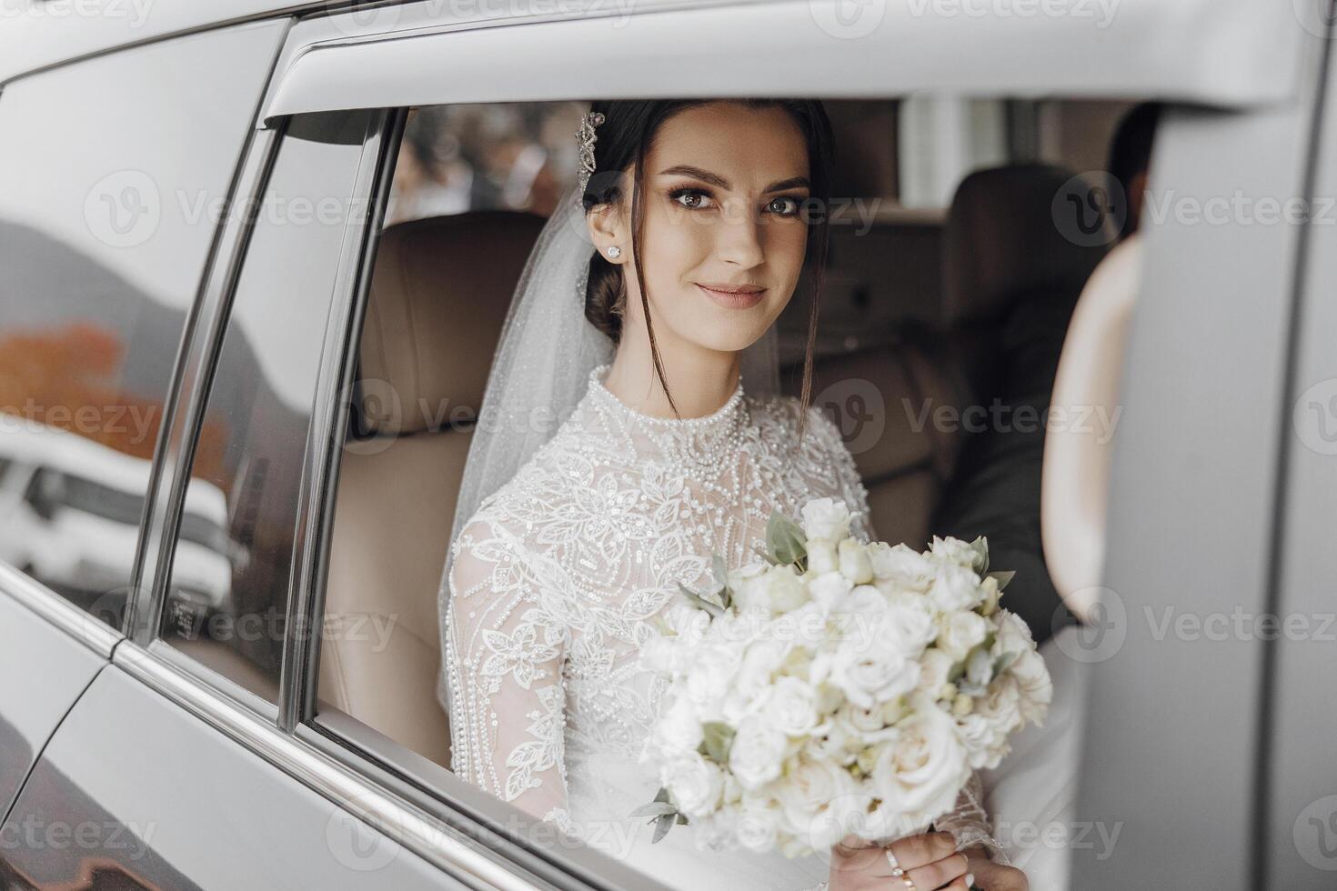 A bride is sitting in a car with a bouquet of white flowers. She is smiling and looking out the window photo