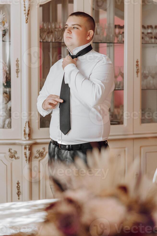A man in a white shirt and black tie is standing in front of a cabinet. He is adjusting his tie and looking at the camera. Concept of formality and attention to detail photo