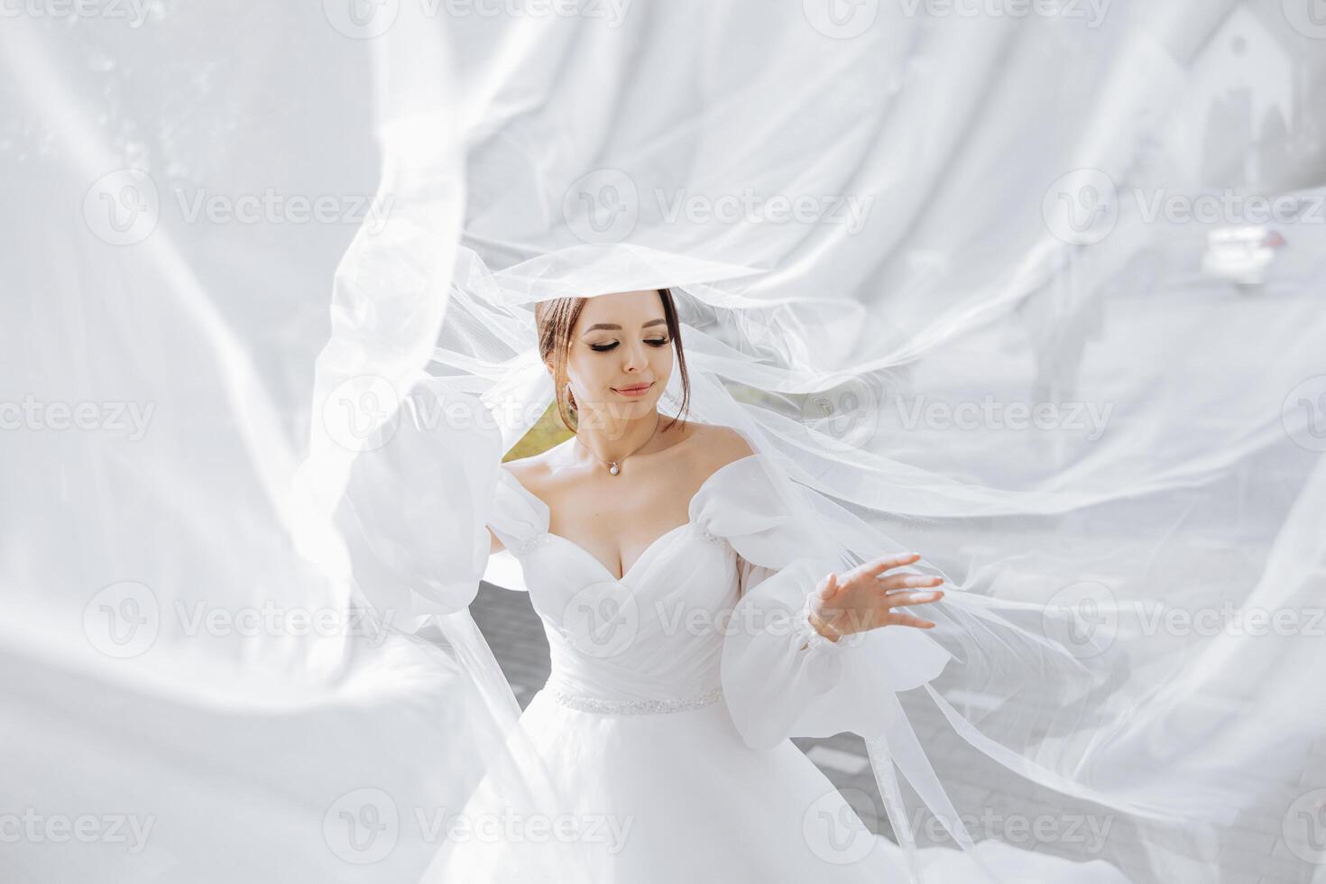 A woman in a white wedding dress is standing in front of a white curtain. She is smiling and she is happy photo