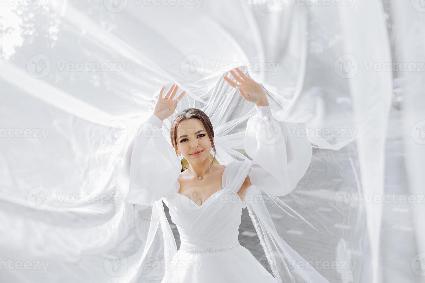 A woman in a white wedding dress is standing in front of a white curtain. She is smiling and she is happy photo