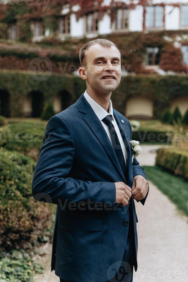 A man in a blue suit and white shirt is standing in front of a building. He is wearing a tie and a white flower in his pocket photo