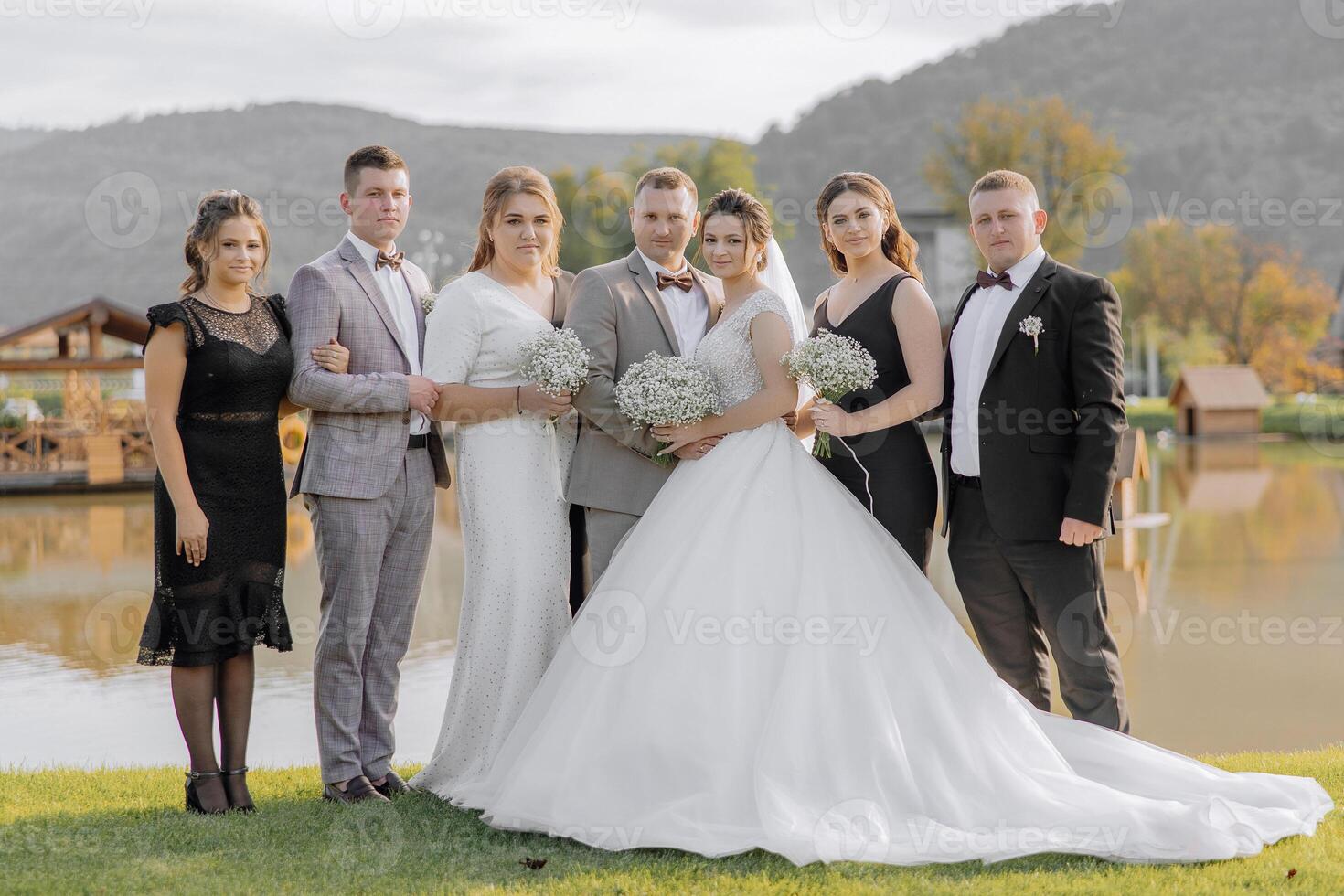 A group of people are posing for a photo, including a bride and groom. The bride is wearing a white dress and the groom is wearing a suit. The group is standing in front of a body of water photo