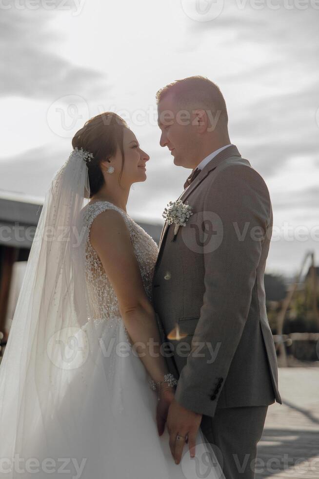A bride and groom are standing together, smiling and hugging. The bride is wearing a white dress and the groom is wearing a suit photo