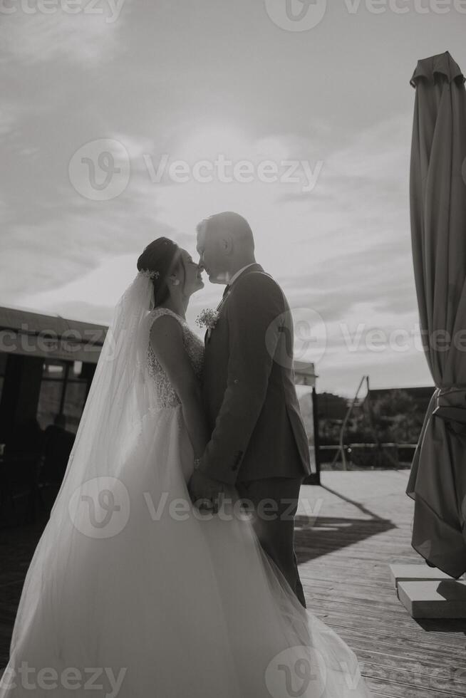 A bride and groom are kissing each other on a wooden deck. The bride is wearing a white dress and the groom is wearing a suit. The scene is romantic and intimate photo