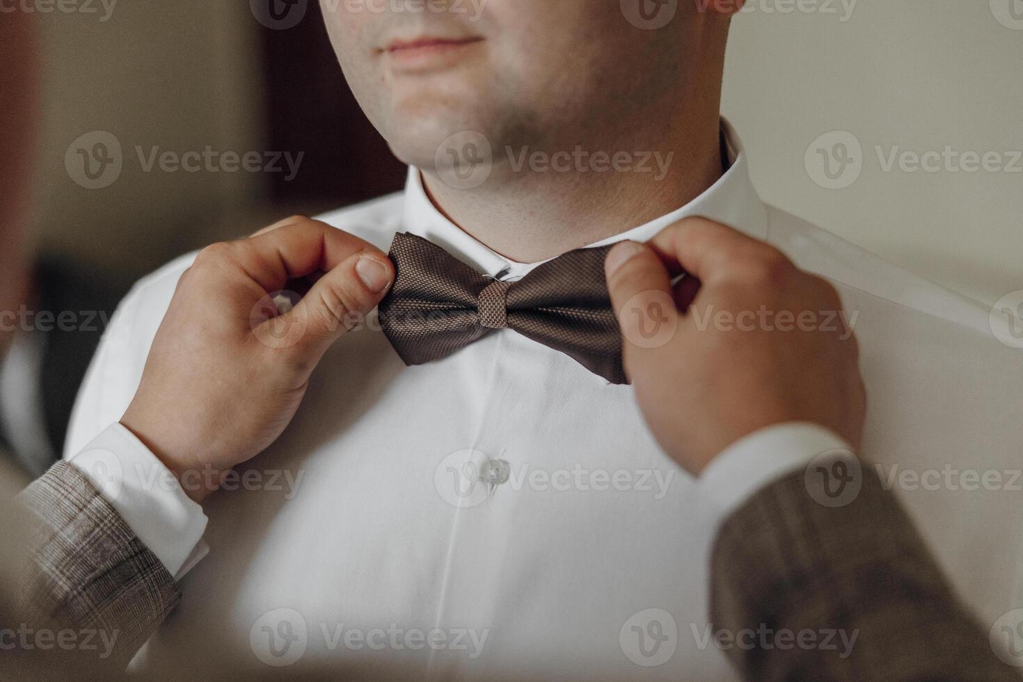 A man is getting ready to wear a bow tie. A man is helping him tie it. The man is wearing a white shirt photo
