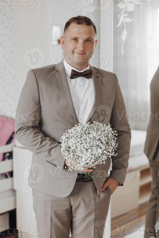 A man in a suit holding a bouquet of white flowers. He is smiling and looking at the camera photo