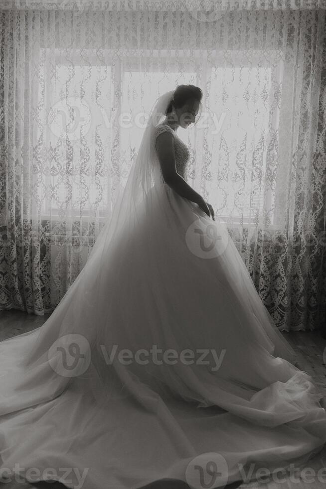 A woman in a wedding dress stands in front of a window. The dress is white and the woman is wearing a veil. The image has a romantic and elegant mood photo