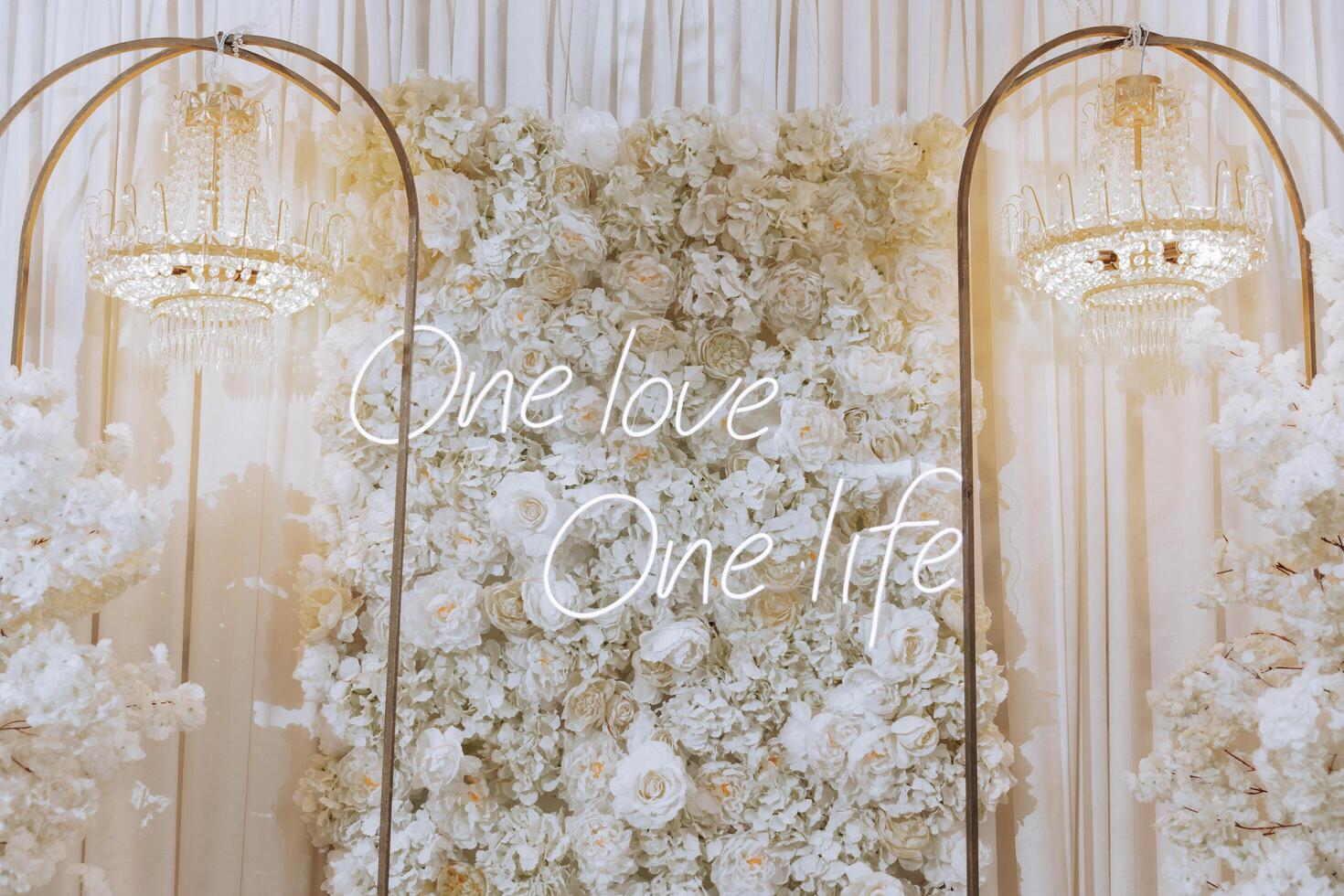 A white floral wall with a neon sign that reads The sign is illuminated and stands out against the white background. Scene is one of love, unity, and positivity photo